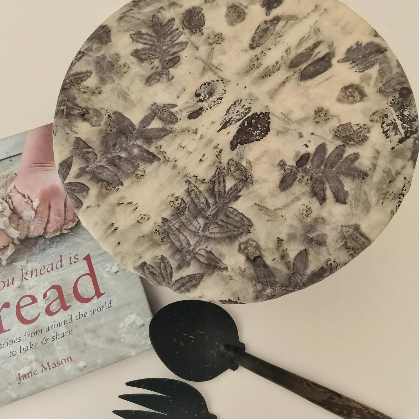 Charlotte couvre-plat en tissu ancien teint avec des plantes sauvages, posée sur un plat rond. Impression végétale en nuances naturelles, accompagnée d’un livre de recettes et de couverts artisanaux.