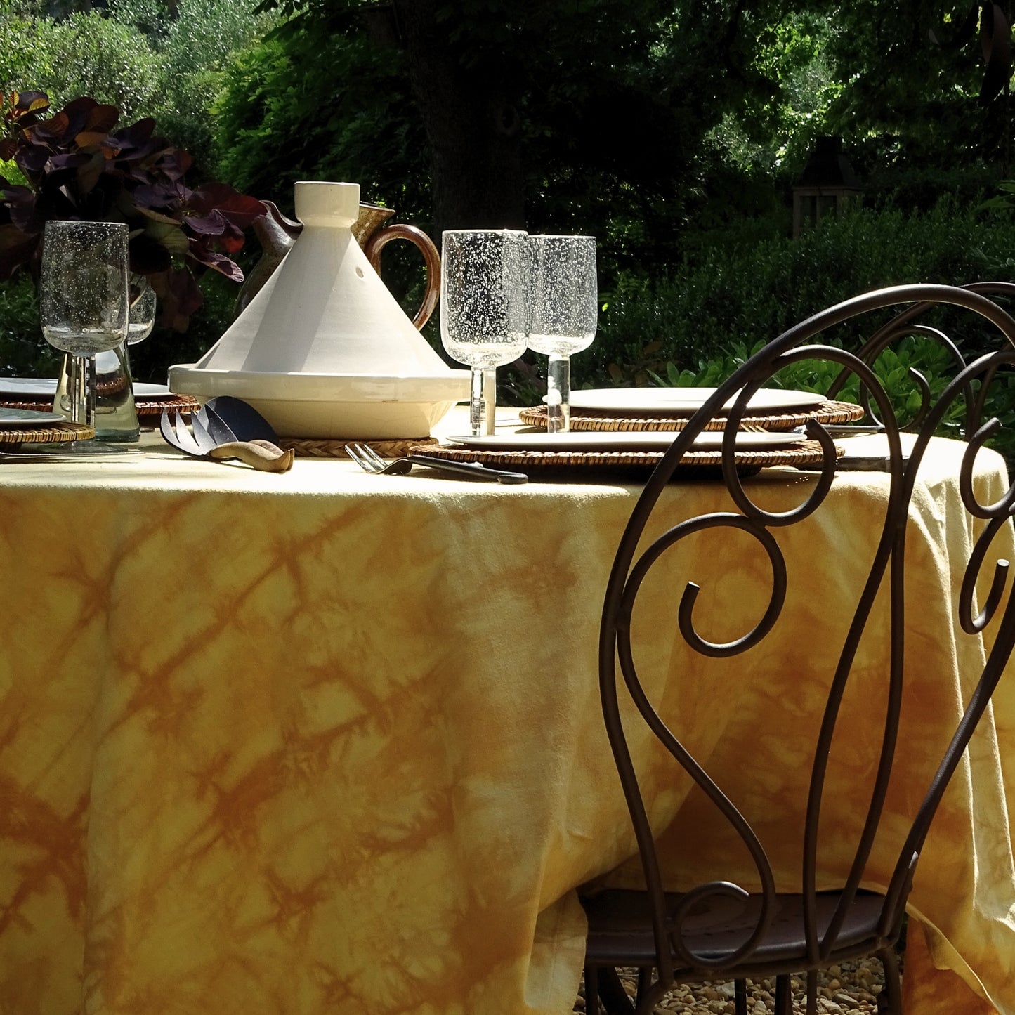 Nappe carrée en coton ancien, aux tons jaunes tie & dye, posée sur une table en extérieur. Pièce unique réalisée en teinture végétale avec pelures d’oignons rouges et jaunes.