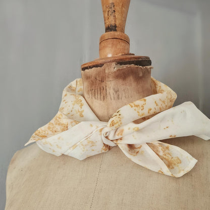 Foulard carré en coton teint naturellement, noué autour du cou d’un buste ancien. Motifs végétaux ocre sur fond blanc cassé, mis en valeur par une lumière douce et un fond neutre.