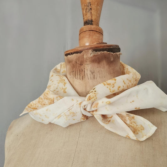 Foulard carré en coton teint naturellement, noué autour du cou d’un buste ancien. Motifs végétaux ocre sur fond blanc cassé, mis en valeur par une lumière douce et un fond neutre.