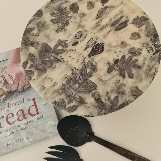 Charlotte couvre-plat en tissu ancien teint avec des plantes sauvages, posée sur un plat rond. Impression végétale en nuances naturelles, accompagnée d’un livre de recettes et de couverts artisanaux.