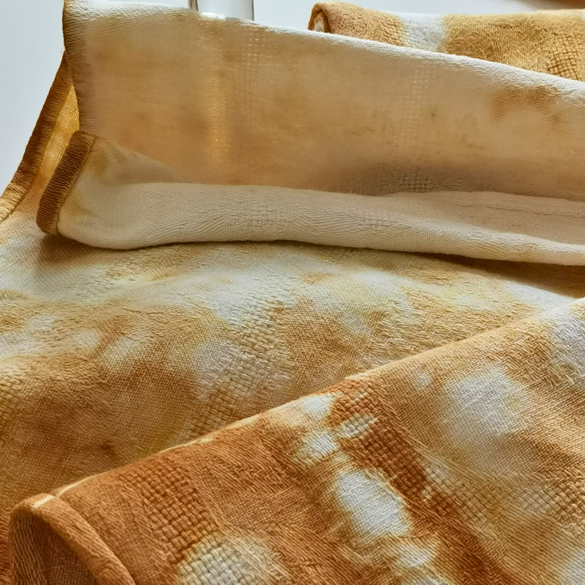 Gros plan sur des serviettes en lin anciennes, teintées naturellement avec des nuances de jaune doré et ocre. Le tissu texturé révèle des motifs tie and dye uniques, mettant en valeur un travail artisanal et des irrégularités naturelles. Une composition douce et chaleureuse qui évoque l’authenticité et le charme du fait main.