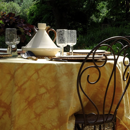 Nappe carrée en coton ancien, aux tons jaunes tie & dye, posée sur une table en extérieur. Pièce unique réalisée en teinture végétale avec pelures d’oignons rouges et jaunes.