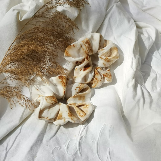Chouchous en soie blanche avec motifs brun clair, teints naturellement, posés sur un drap blanc avec des pampas séchées.
